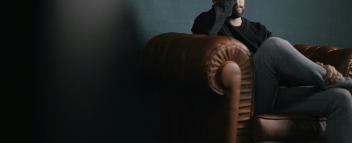 Darkly lit photograph of a man sat on a leather sofa. The man has his head in his hand and is thinking.