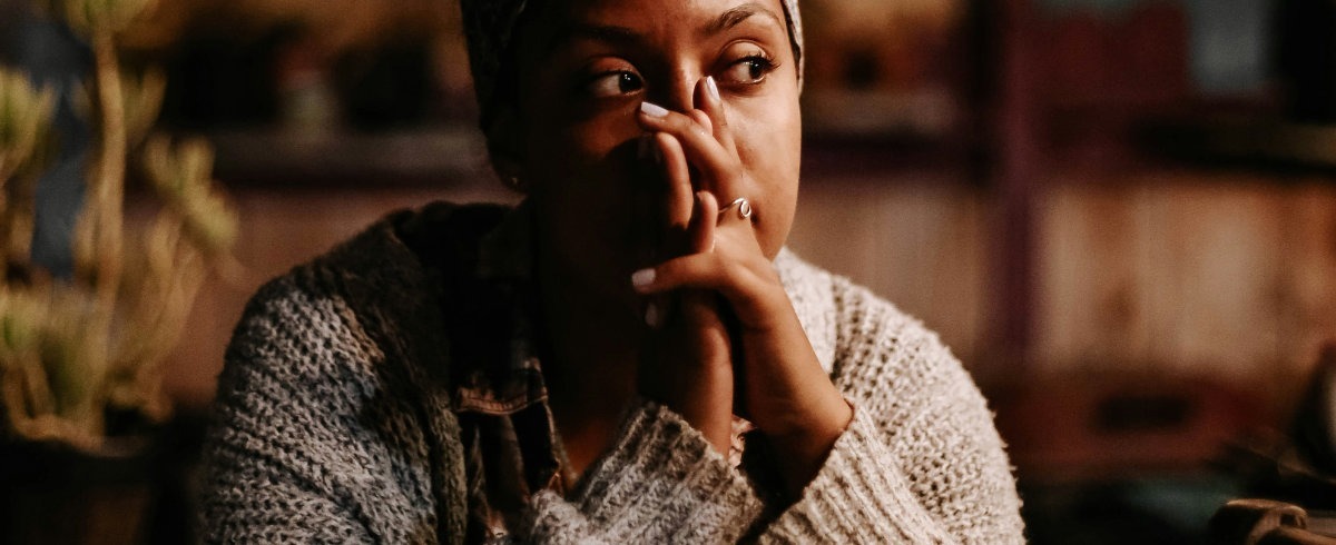 Darkly lit photograph of a woman in her 30's sat at a table. She is looking into the middle distance and contemplating something.