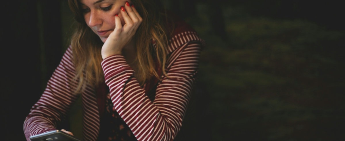 Photograph of a woman sat at a table. She is wearing a striped top and is looking at her mobile phone. She seems absorbed or reflecting on the message she has received.